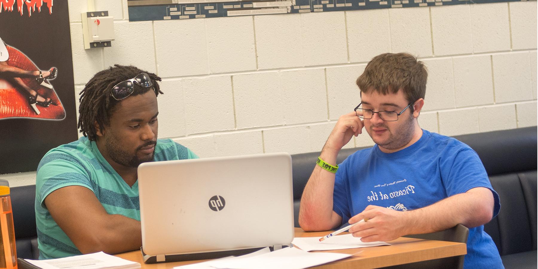 Two students work together in a room with white brick walls and blue benches. The wooden table they are at is covered in papers, with an open HP laptop centered in the frame. They appear to be studying together.